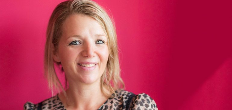 The author Clover Stroud, a woman with blue eyes and blonde hair, wearing a leopard print top and standing against a pink background.