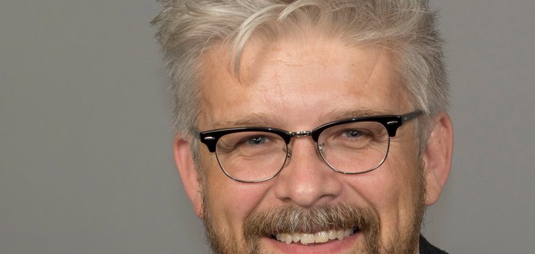 Simon Kovesi looks straight at the camera, with a grey hair, beard and moustache. You can only see his head and shoulders, he is wearing glasses and he is smiling.