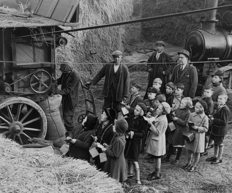 A group of schoolchildren watching a threshing machine in action. The image is in black and white and is from during World War Two.