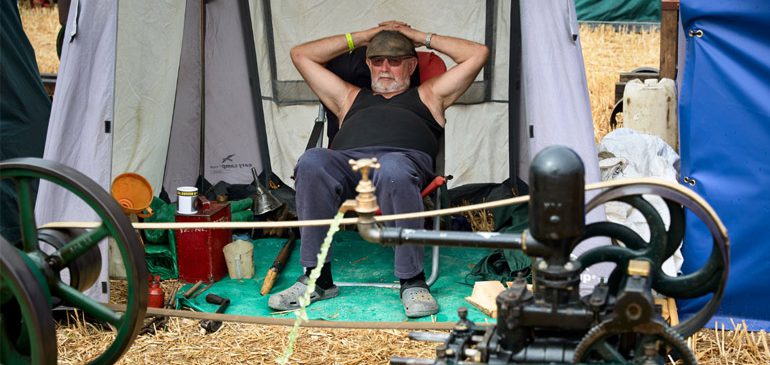 A photograph of a man in a black tank top with sunglasses and a flat cap, with his hands behind his head. In the foreground an open tap covers his crotch.