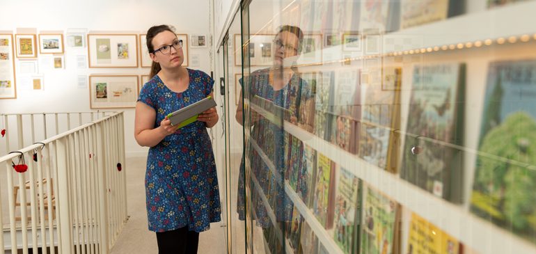 Researcher holding an ipad and looking closely at the wall of books in the Ladybird Gallery at the MERL