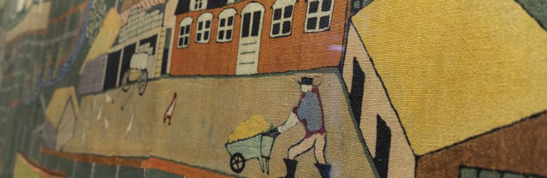 Detail of textile wall-hanging of Kent, England, with dyed decoration showing a figure with a wheelbarrow, some chickens, and a water barrow in a traditional farmyard
