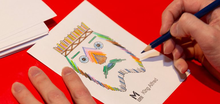 Close up of a hand holding a pencil to colour in a line drawing of the face of the MERL King Alfred