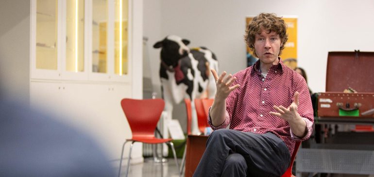 Pete Flood sitting and gesturing whilst speaking with MERL cow in background
