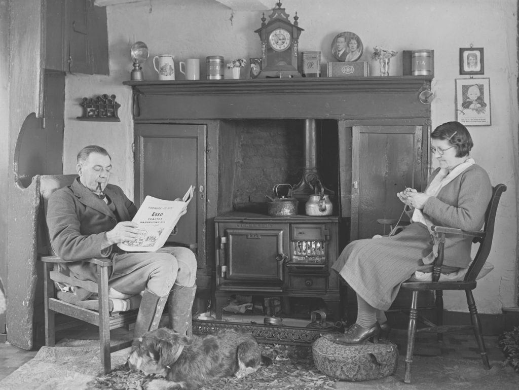 A couple reading in a cottage by a fireplace.