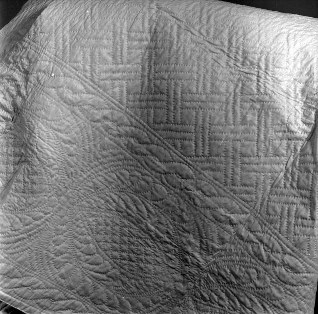 White wholecloth quilted bedcover made of linen. It is quilted with floral and geometric designs in running stitch.