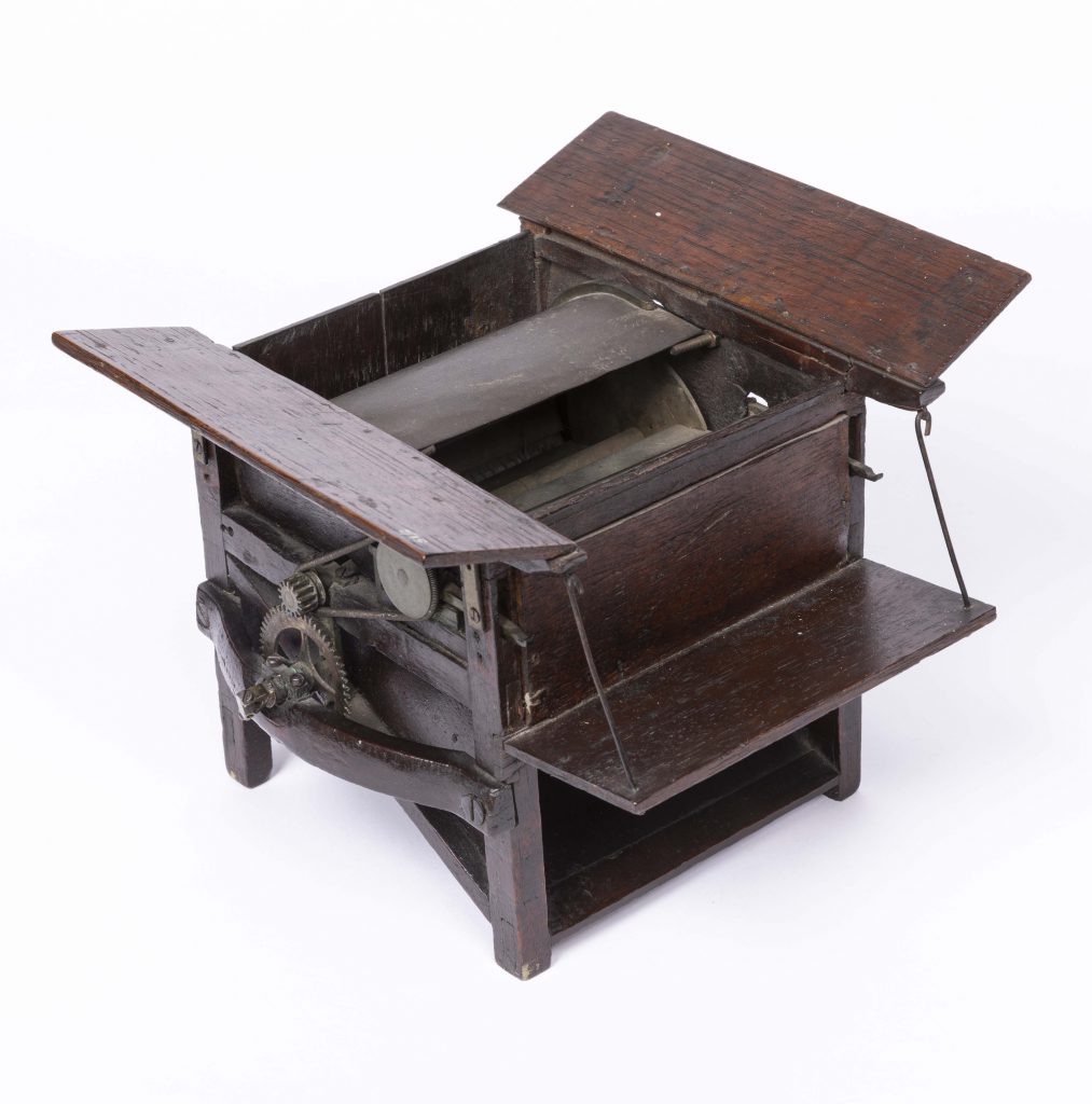 Small dark wooden threshing machine mechanism with cogs and workings visible to one side and open 'flaps' at the top.