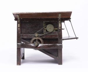 Small dark wooden threshing machine mechanism with cogs and workings visible to one side and open 'flaps' at the top.