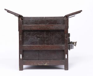 Small dark wooden threshing machine mechanism with cogs and workings visible to one side and open 'flaps' at the top.