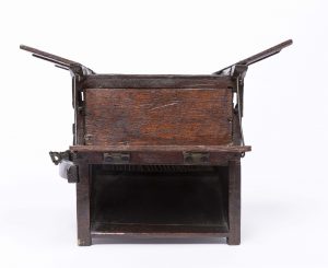 Small dark wooden threshing machine mechanism with cogs and workings visible to one side and open 'flaps' at the top.