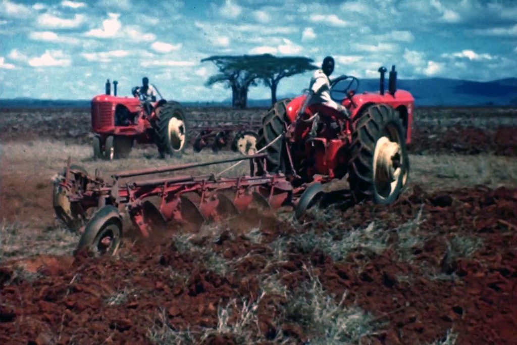 Two workers drive tractors and cultivate the soil with acacia tress on the horizon in the background