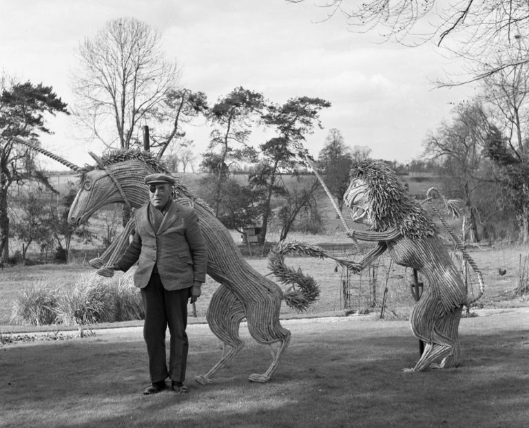 Black and white photograph of a man with an eye patch and cap standing beside large straw sculptures of a lion and a unicorn, with trees in the background.