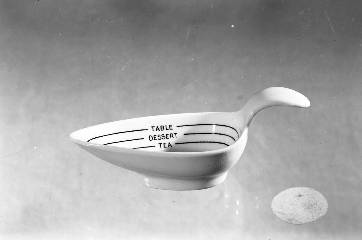 This is a porcelain measuring spoon used when cooking or for measuring medicine. It is white, with 'tea', 'dessert' and 'table' marked around the inside.
