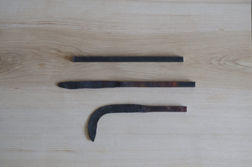 Three roughly finished dark coloured steel bars showing three stages of forging a curved blade, all sitting on a table top surface.