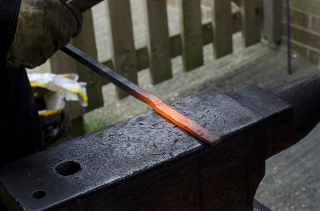 Image of hot metal resting on an anvil and in teh process of being beaten into a flat blade shape. A wooden fence and brick wall are visible in the background.