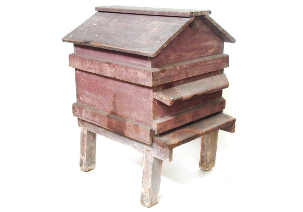 A wooden beehive with a sloping roof, resting on a wooden base with legs, some of which seem to be slightly squint.