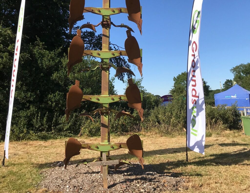 A plough buried upright with a small wooden cross in front of it, with trees in the background and a banner to one side that reads 'Groundswell'