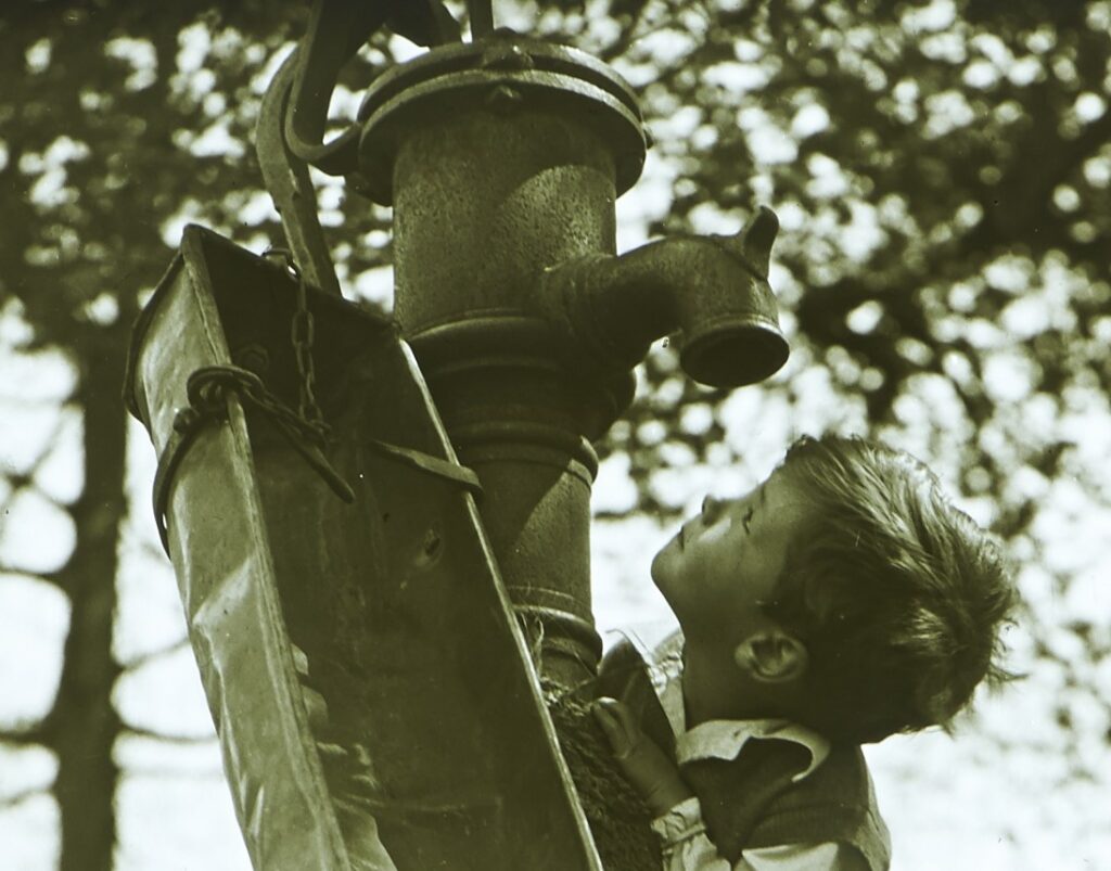 Detail of a young boy looking at a village water pump with a tree in the background.