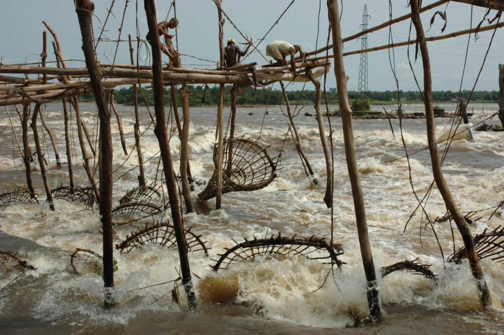 Fishers balance on a wooden structure over crashing waves, dangling a series of conical wooden fish traps into the water.