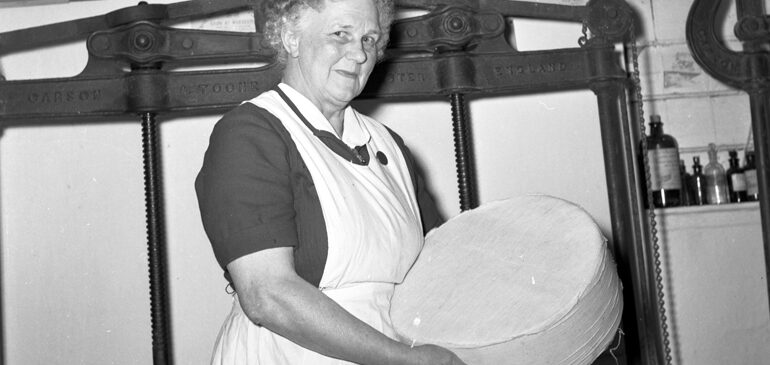 A black and white photo of lady in an apron holding a large round of double Gloucester cheese