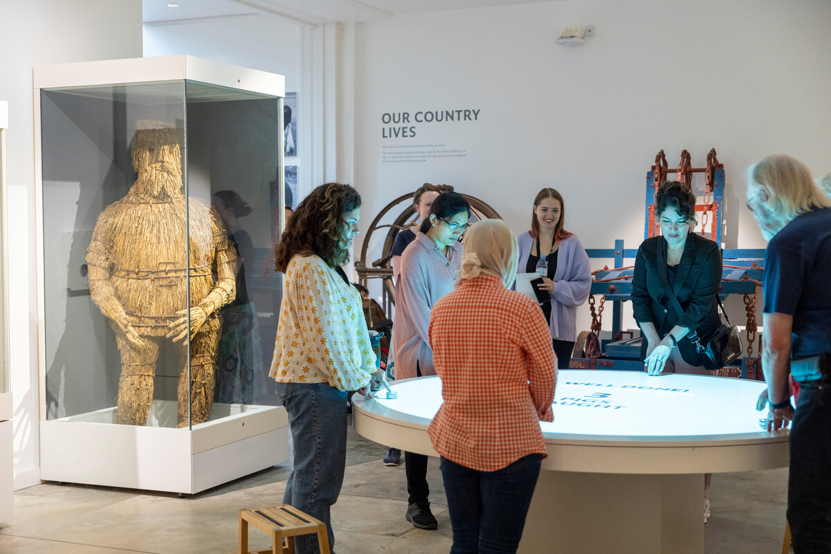 a group of people stand around a round table with a game projected onto it from above in the Our Country Lives gallery at The MERL