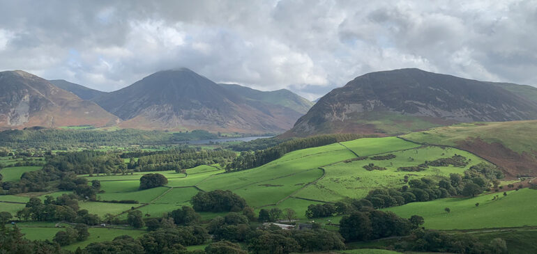 A photograph of the a Lake District landscape with green fields and trees in the foreground and fells in the distance