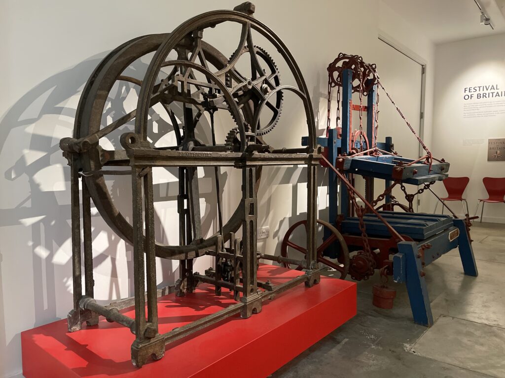 Well-head on a plinth beside a hay press, on display in a gallery