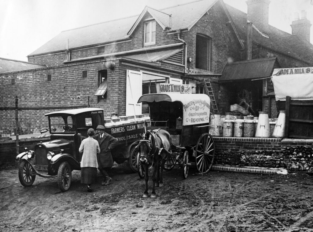 Black and white photo showing a horse and cart loaded with milk bottles and an old motorcar with 2 people having a conversation, with a brick building in the background