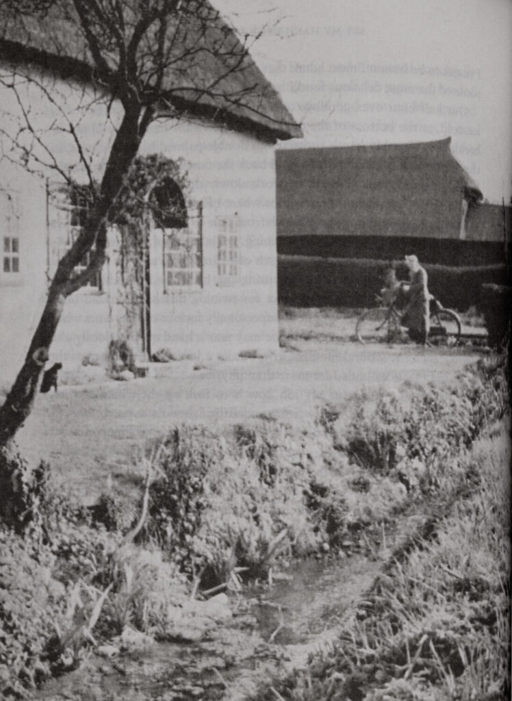 Black and white photograph depicting a cottage, stream, and a figure coming home on a bicycle.