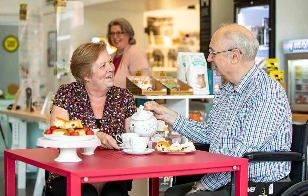 A man and woman make eye contact and smile while enjoying cream tea - complete with fine china, scones and strawberries - in The Museum of English Rural Life café