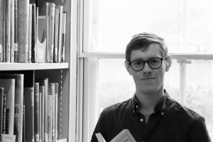 A black and white photo of poet in residence Jack Thacker standing in front of a bright window next to a bookcase, holding a book.