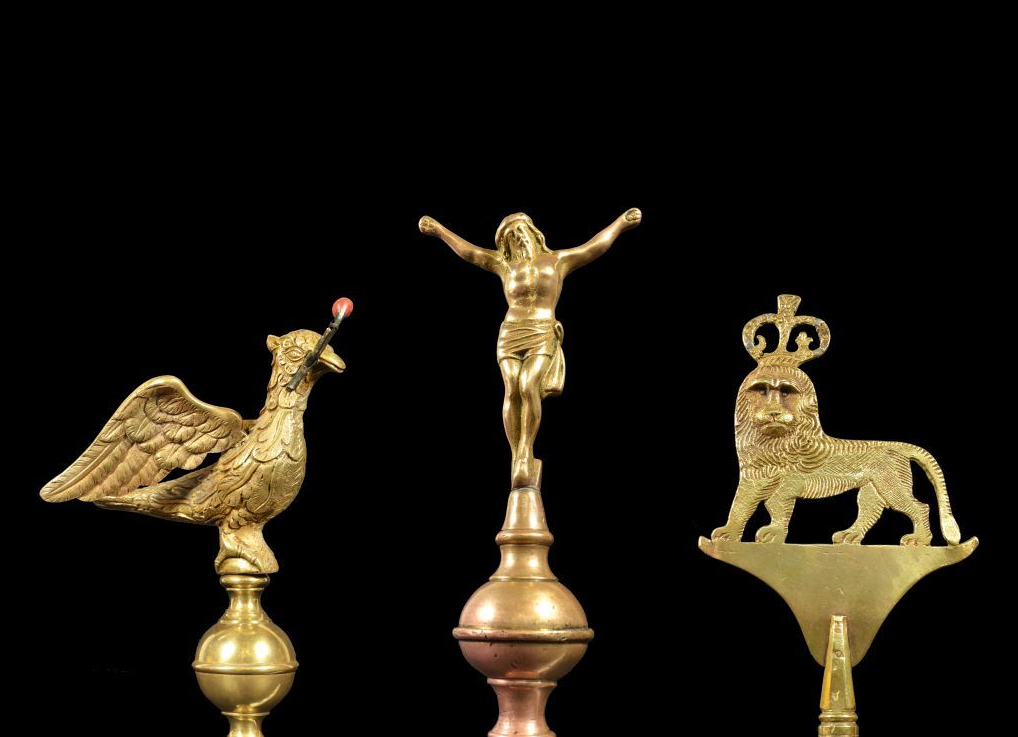 Three brass poleheads against a black background. On left is a bird holding a flaming torch, in the middle a crucified Jesus and on the right a crowned lion.