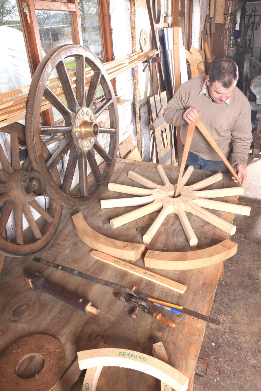 A man measuring the distance between spokes for a wooden wagon wheel, which lies on a table half-finished.