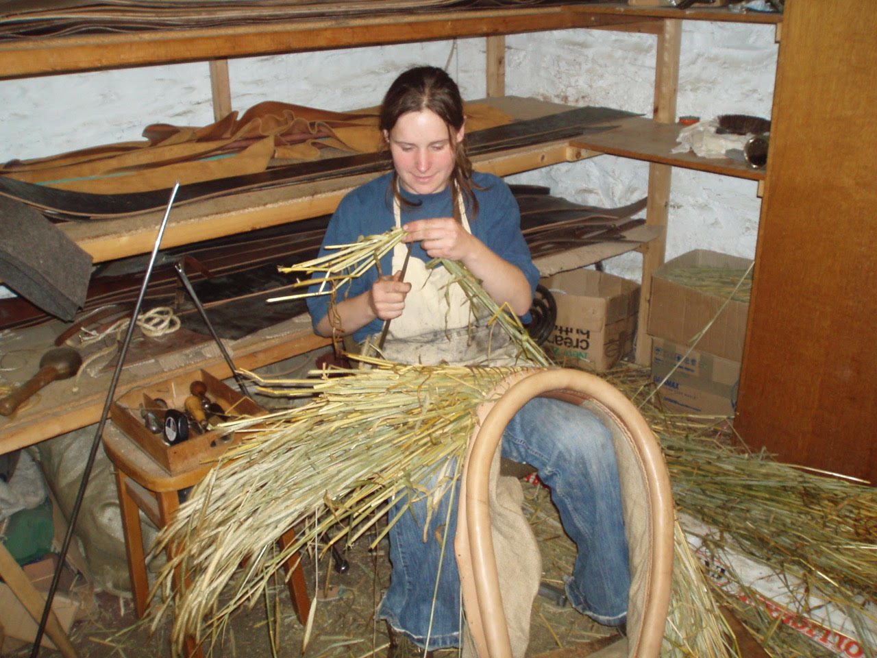 A woman in a blue shirt, white apron and blue jeans sat down, stuffing a leather horse collar with straw.
