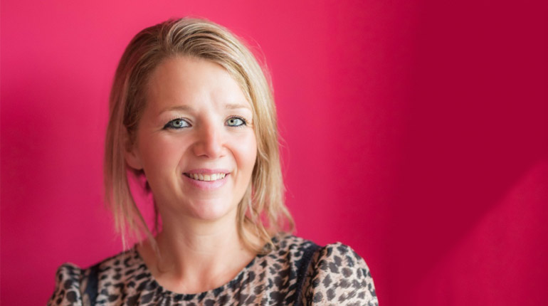 The author Clover Stroud, a woman with blue eyes and blonde hair, wearing a leopard print top and standing against a pink background.