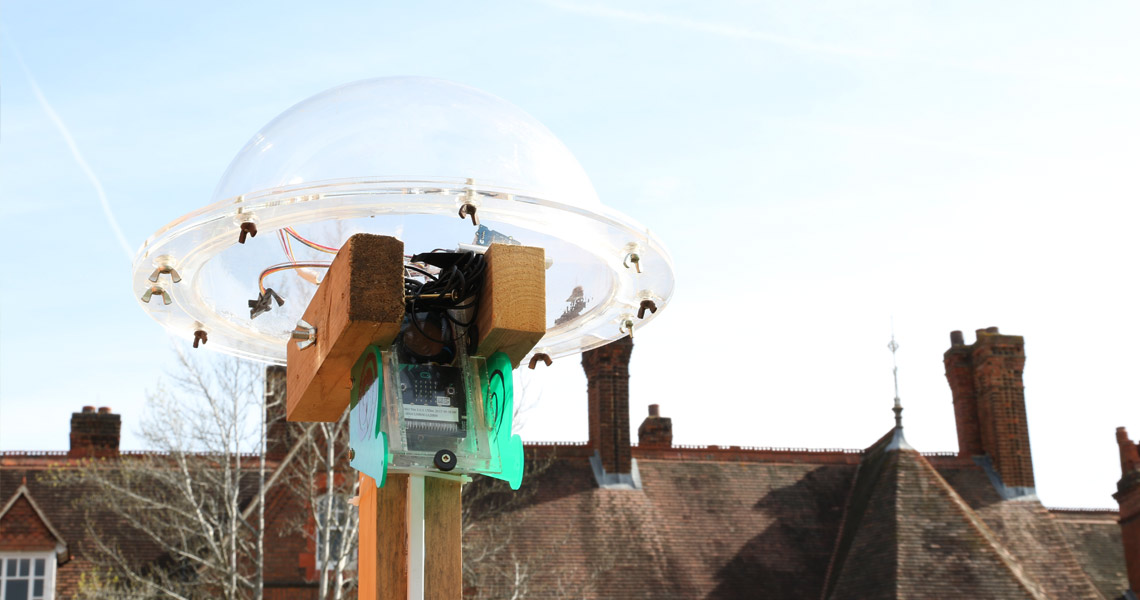 A plastic dome on top of a wooden pole, housing a BBC MicroBit. The roof of the building is in the background against a blue sky.