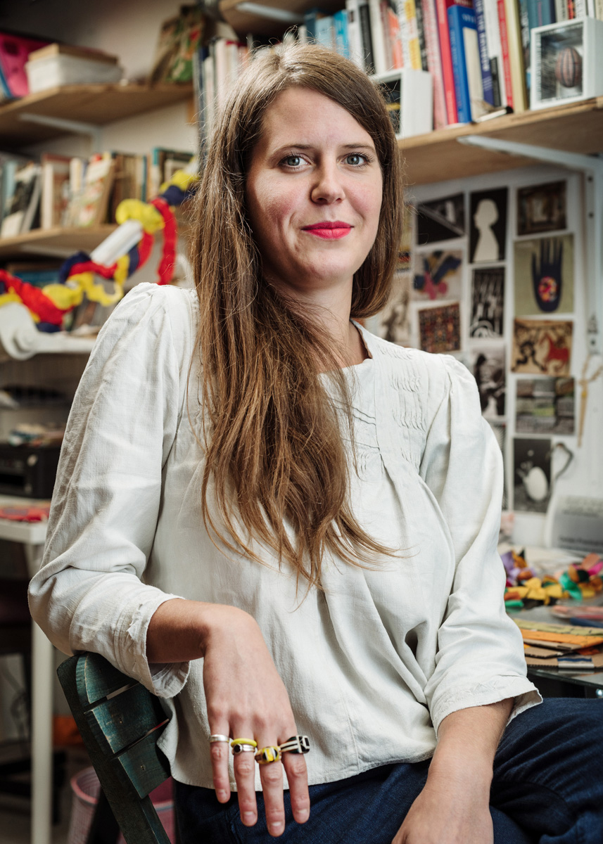 Caitlin Hinshelwood sits on a chair facing the camera, with an arm draped on the back of the chair. She has long brown hair, is white and is wearing a white shirt. Behind her is her artist studio.