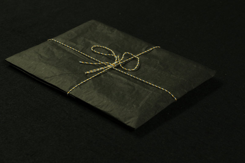 A small square package wrapped in black tissue paper and tied with a multicoloured string sits on a black surface.