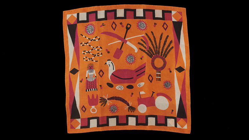 An orange scarf covered in rural designs, such as a tractor and a corn dolly, sits on a black background.