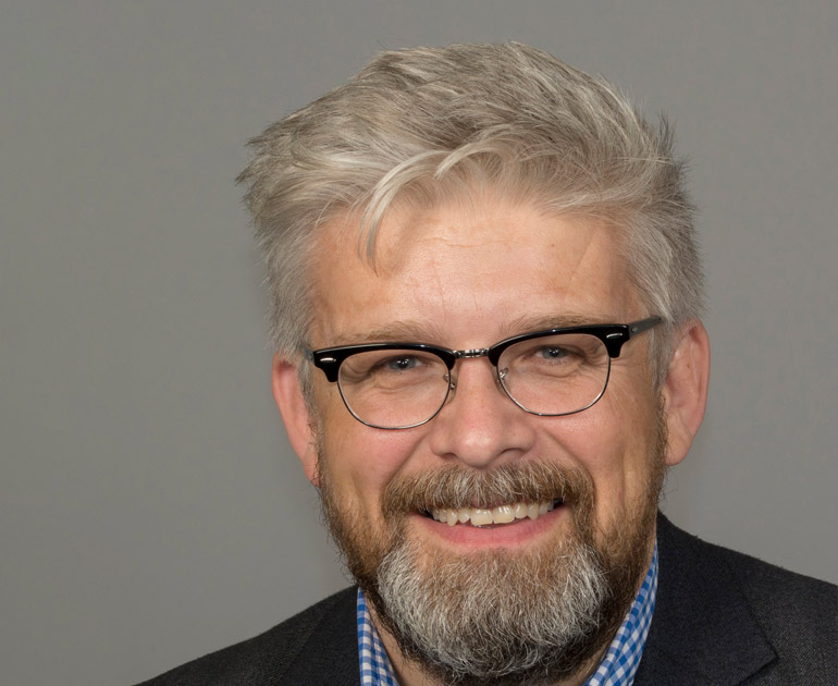 Simon Kovesi looks straight at the camera, with a grey hair, beard and moustache. You can only see his head and shoulders, he is wearing glasses and he is smiling.