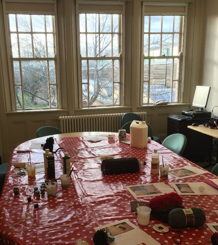 A table in a room with three windows. The table is covered in craft materials