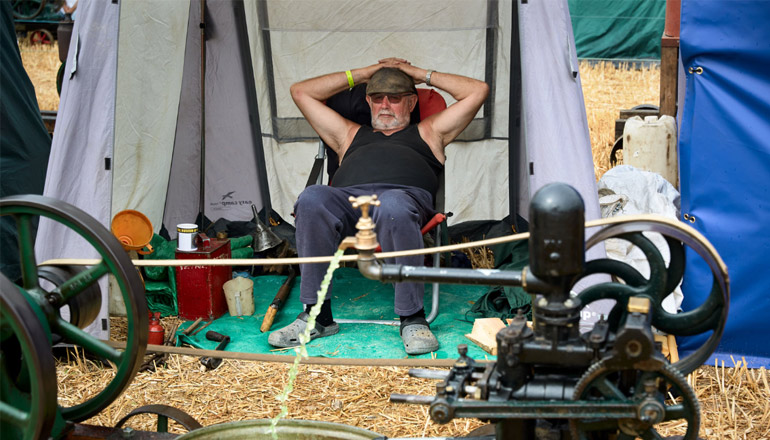 A photograph of a man in a black tank top with sunglasses and a flat cap, with his hands behind his head. In the foreground an open tap covers his crotch.