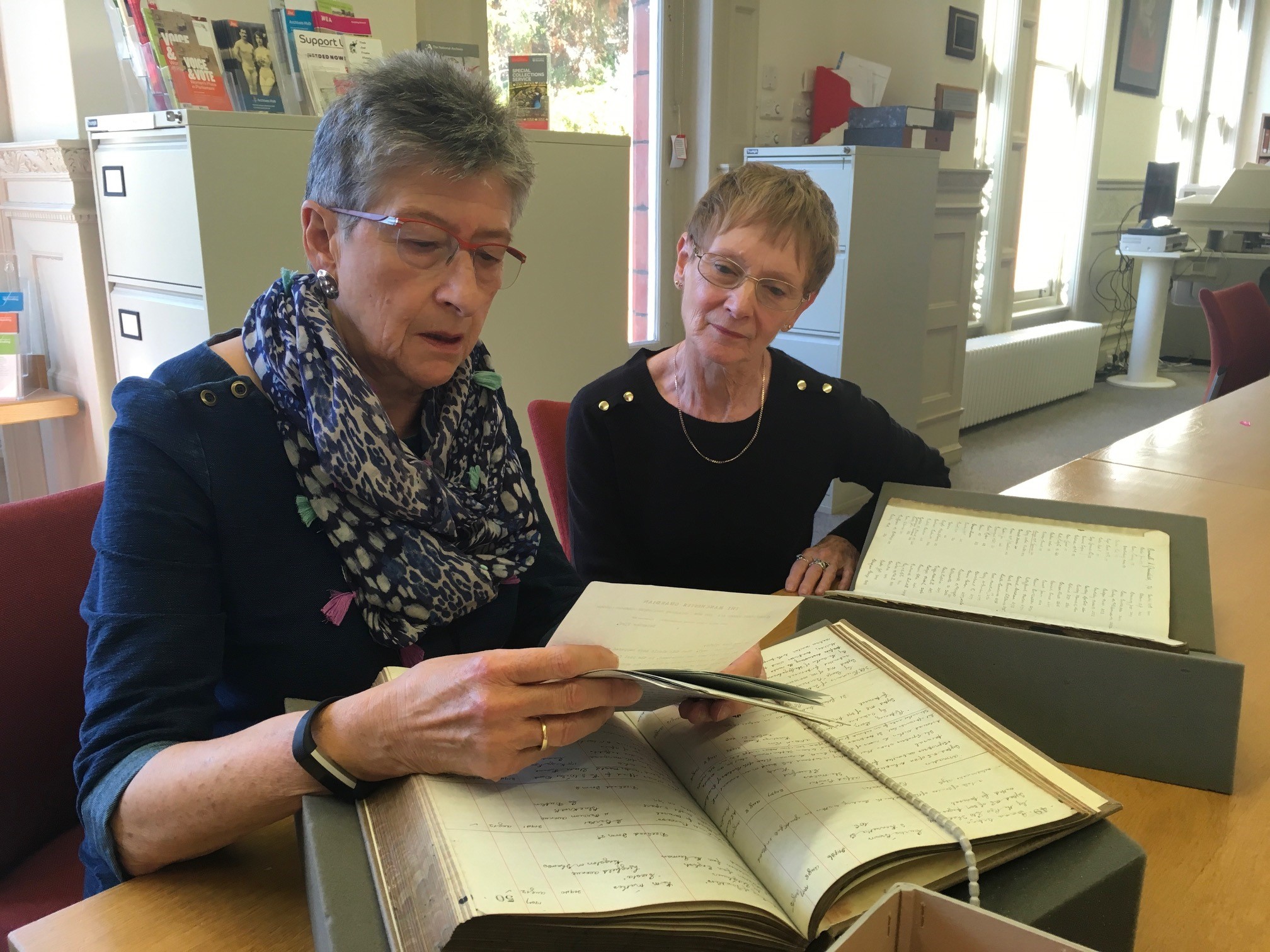 Two female volunteers sitting at the large table in the reading room with historic library books resting on foam and looking at letters