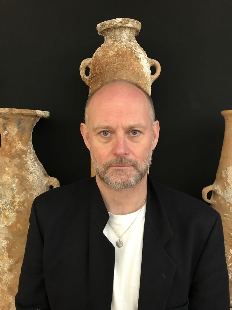 The artist Steven Claydon looking into the camera against a black background with ancient pots behind him