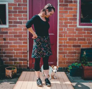 Hannah James standing on a wooden platform wearing black clogs and looking down at the small dog by her feet