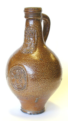 A Bellarmine jug, with a narrow base, wide centre, and narrow top - The top is more narrow than the base. The jug is a mid-brown colour, and has a mottled glaze. The top of the jug is ridged with three bands. The Jug has a handle on the reverse, the top of which sits at the top of the jug, and finishes about 6 cm down the body. On the front of the jug is engraved a face of a bearded man, below which is a circular motif in the centre of which is a lion-type creature.