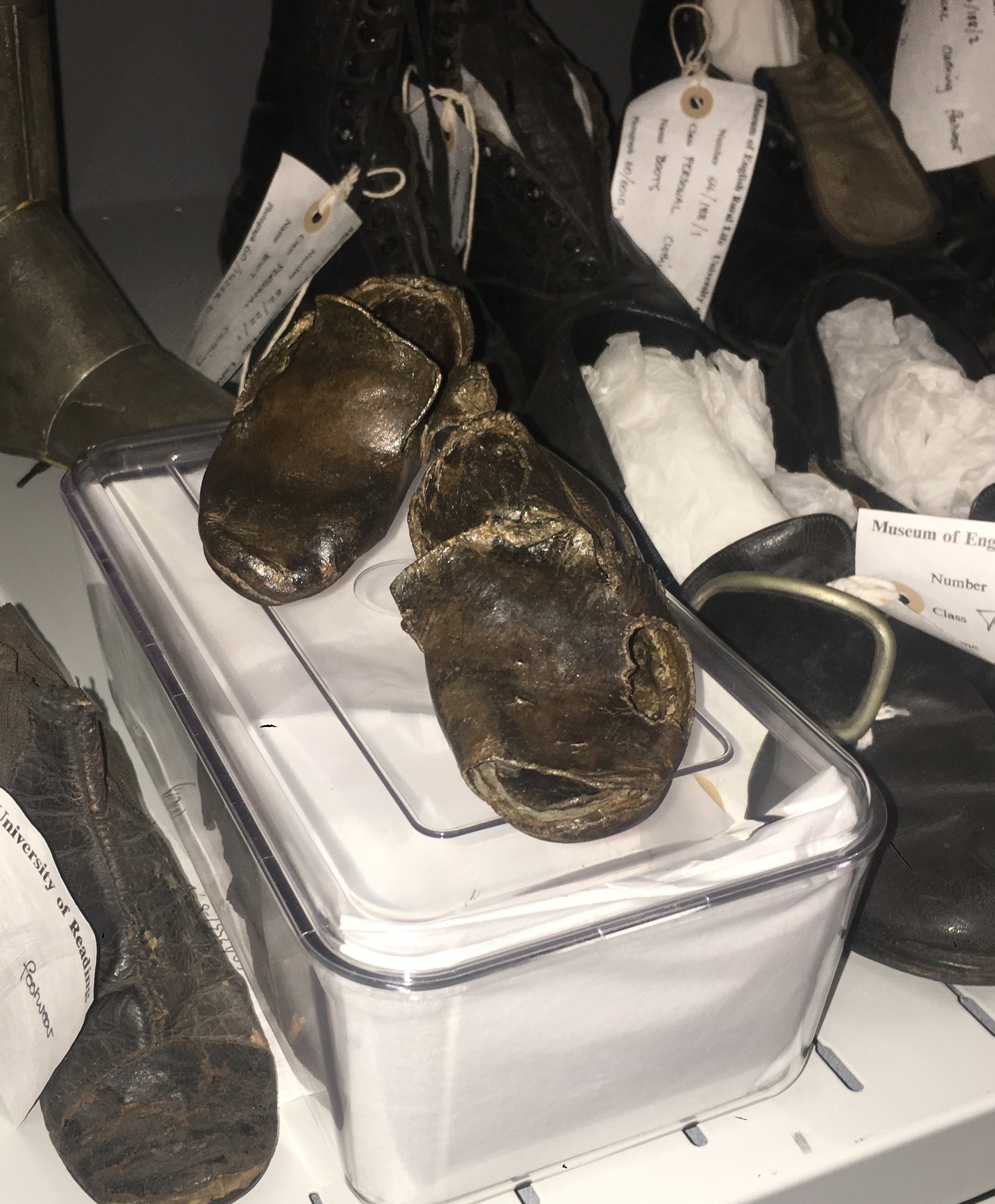 A pair of brown children's leather shoes, both measuring approx. 8cm in length. Both have been conserved and are, as a result, slightly shiny. One shoes has two holes, one in the toe and one in the side. The other is fairly whole, with a few splits in the sides. There are no visible indication of how they were fastened. 