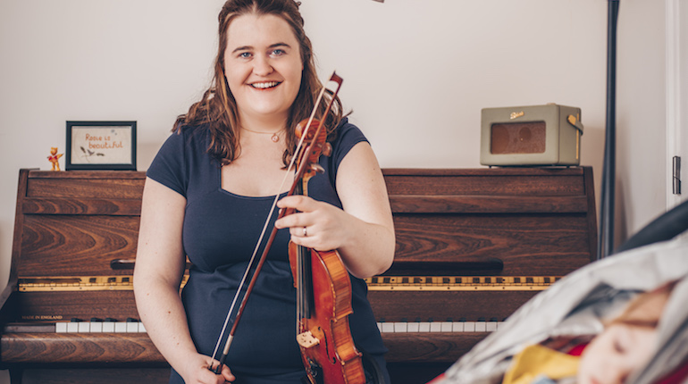 Jackie Oates sitting in front of a piano holding her violin. Her baby is asleep in pushchair in foreground.