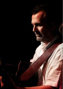 Martin Simpson profile against a black background playing guitar obscured in darkness - to illustrate the seminar at the MERL