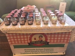 Neat rows of jam jars with red and white gingham lids with open taster jars in front with The Jam Lady logo